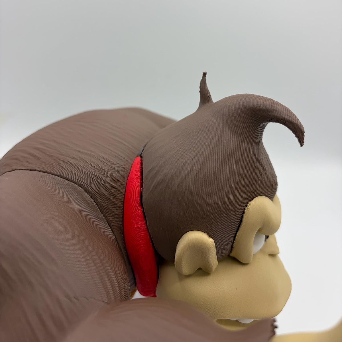 Close-up of a brown monkey figurine or toy with beige face details and a red collar, shown against a plain light gray background.