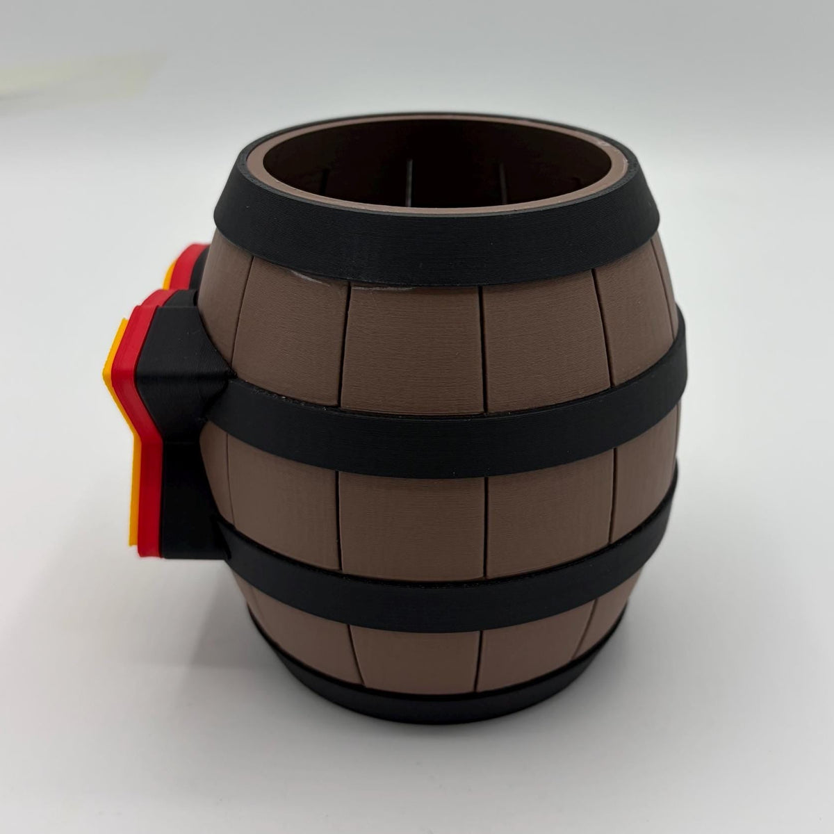 A novelty mug shaped like a miniature brown wooden barrel with black metal bands and a small side handle featuring red and yellow stripes, shown on a light gray studio background.