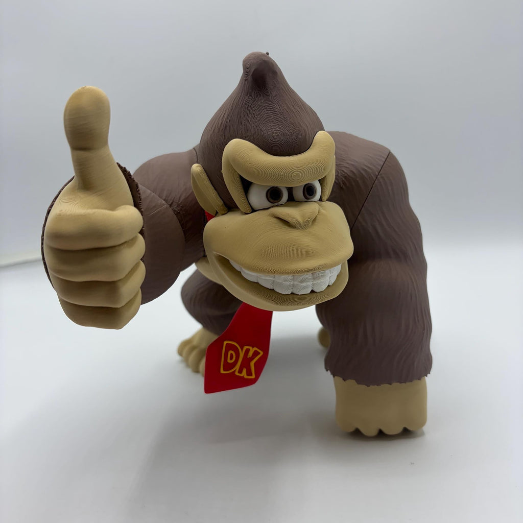 A Donkey Kong action figure or collectible toy standing on a white surface against a light gray background, giving a thumbs-up with its right hand. The figure is brown with tan skin and a red necktie featuring yellow 'DK' lettering.