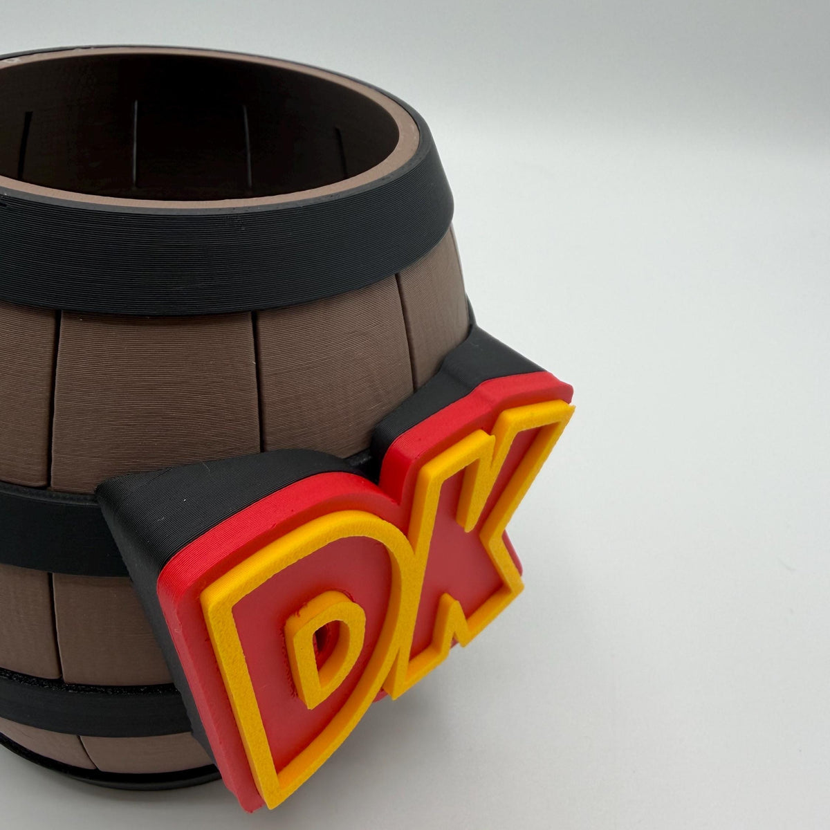 A close-up of a brown barrel-shaped product with black bands and a large red and yellow 'DK' emblem on the side, photographed on a light gray studio background.