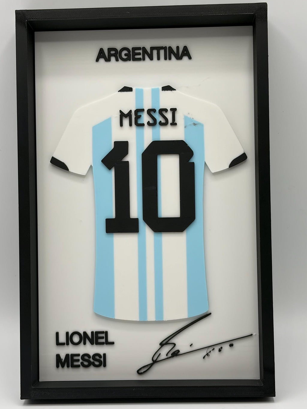 A framed Lionel Messi Argentina jersey display featuring a white and light blue striped soccer shirt with the number 10, black text reading 'ARGENTINA' and 'LIONEL MESSI', and a black signature on a white background inside a black frame.