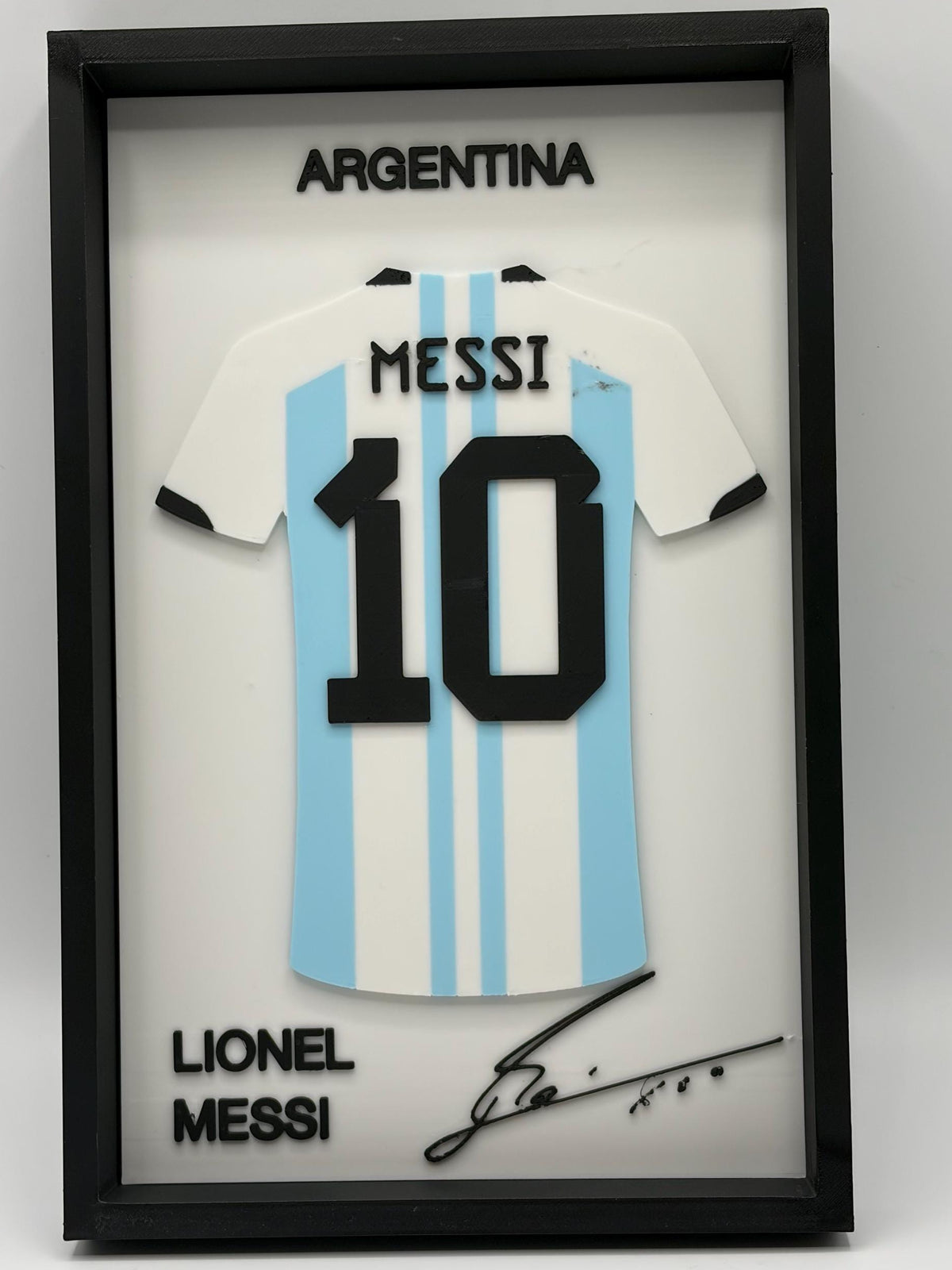 A framed Lionel Messi Argentina jersey display featuring a white and light blue striped soccer shirt with the number 10, black text reading 'ARGENTINA' and 'LIONEL MESSI', and a black signature on a white background inside a black frame.