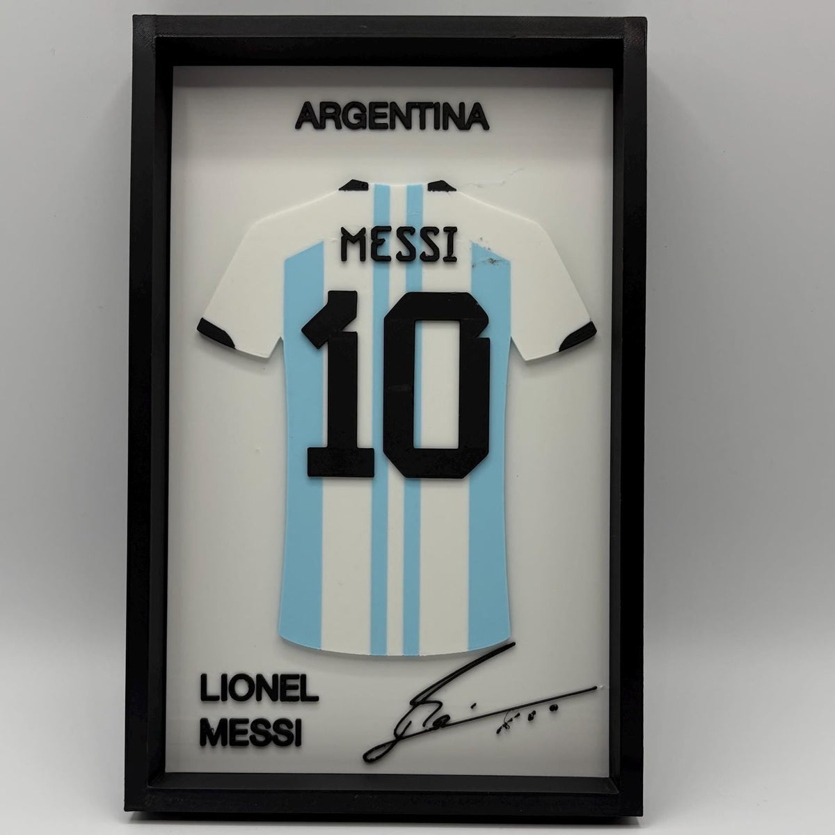 A framed Lionel Messi Argentina number 10 jersey display on a light gray background. The black shadowbox frame contains a white and light blue striped Argentina jersey with 'MESSI' and '10' in black, plus 'ARGENTINA' at the top and 'LIONEL MESSI' with a signature at the bottom.