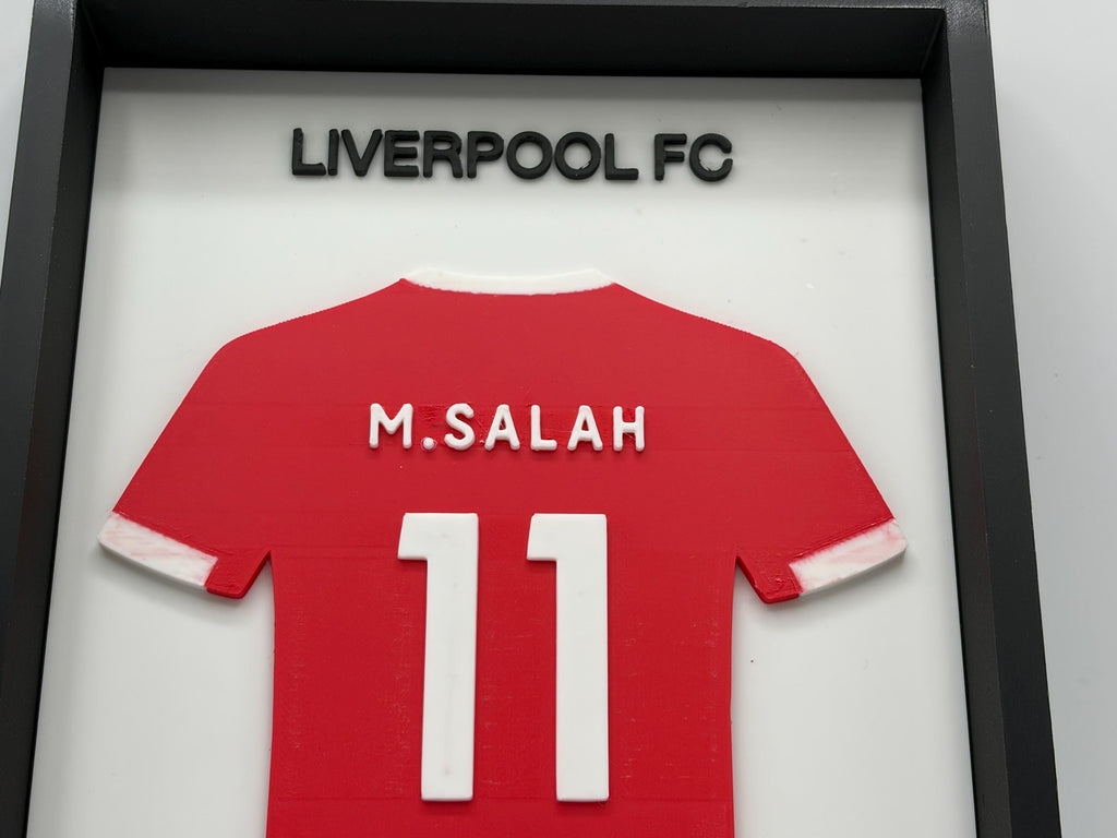 A framed red Liverpool FC jersey with the name M. SALAH and number 11 on the back, mounted inside a black shadowbox frame with Liverpool FC lettering above.