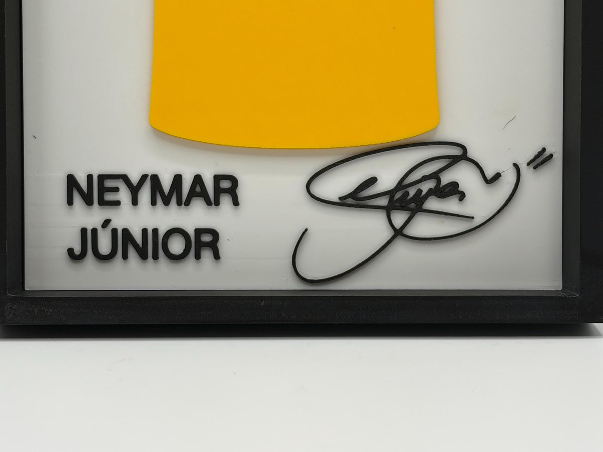 A framed personalized display featuring the text 'NEYMAR JÚNOR' in black, a yellow curved accent at the top, and a black autographed signature on a white background inside a black frame.