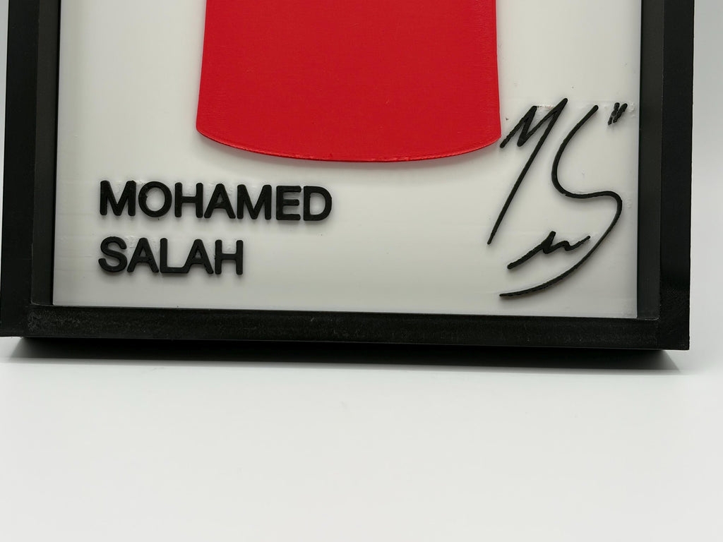 A close-up of a framed personalized wall art piece with a black frame, white background, a red curved shape at the top, the text 'MOHAMED SALAH' in black, and a black signature on the right.