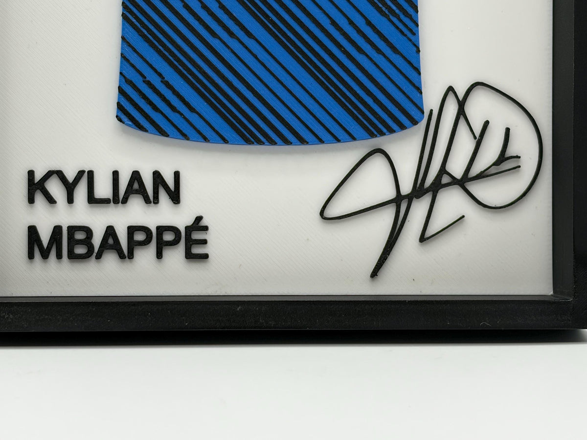 A framed Kylian Mbappé autographed piece featuring a blue and black striped fabric patch, black text reading 'KYLIAN MBAPPÉ', and a black signature on a white background inside a black frame.