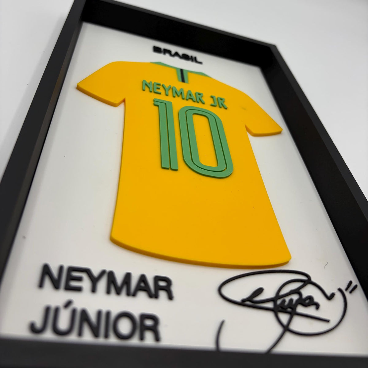A framed custom display featuring a yellow soccer jersey with 'NEYMAR JR 10' and 'BRASIL' in green and black, mounted on a white background with 'NEYMAR JÚNIOR' and a black signature at the bottom.