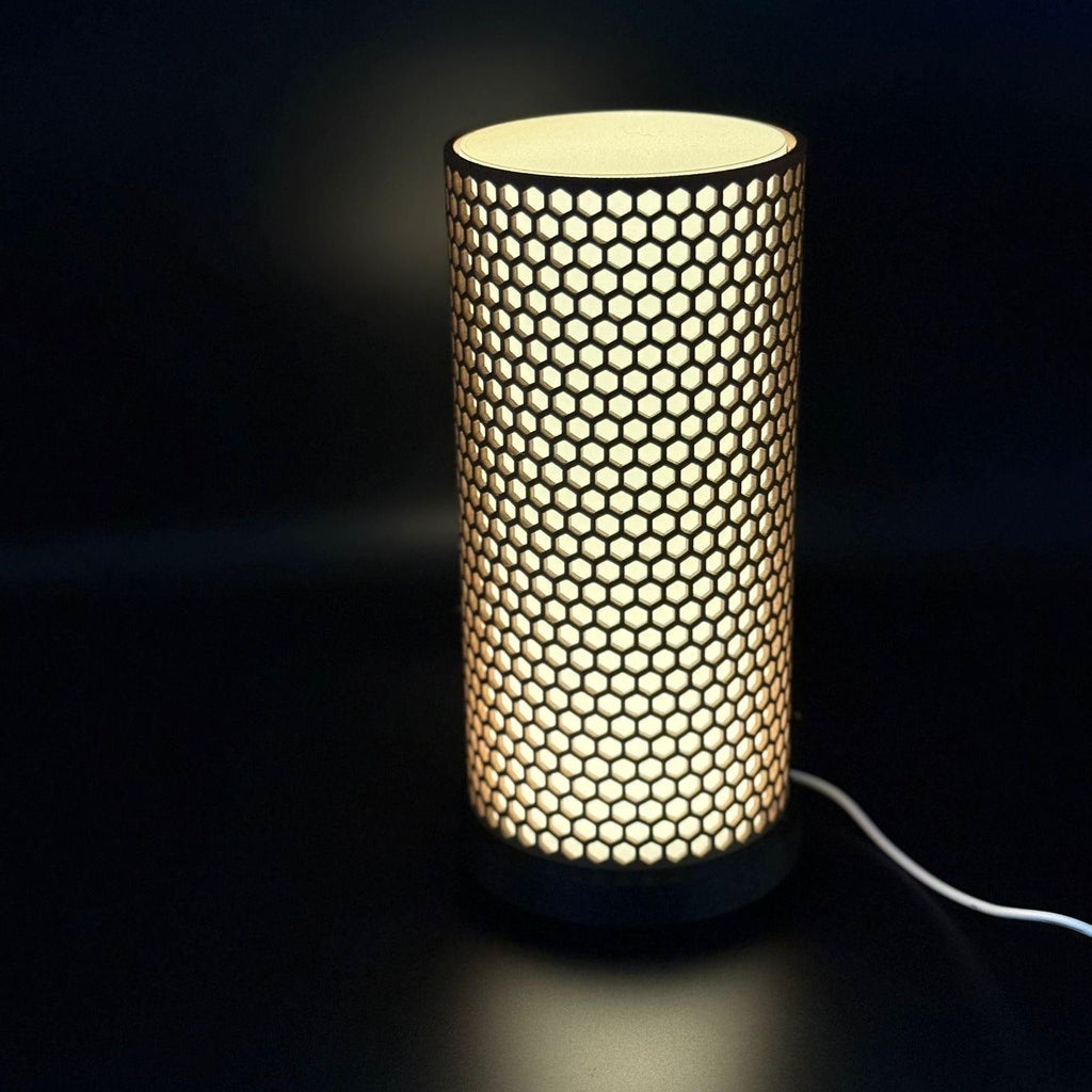 A modern cylindrical table lamp with a glowing honeycomb pattern on a dark surface against a black background.