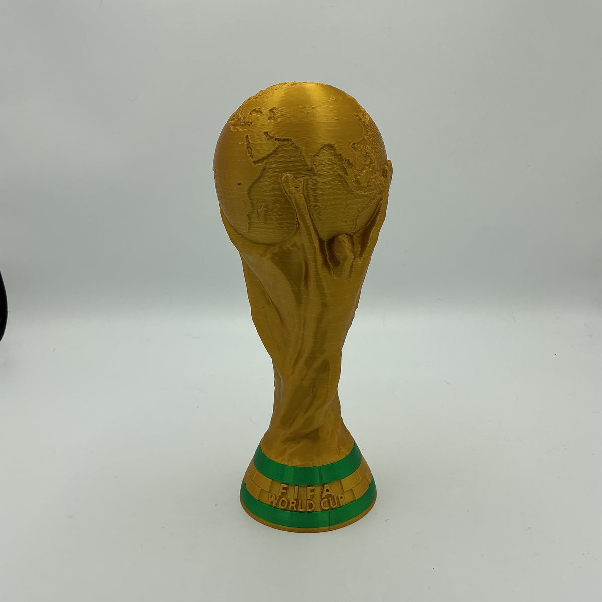 A gold replica of the FIFA World Cup trophy with green accents on the base, standing on a white surface against a light gray background.