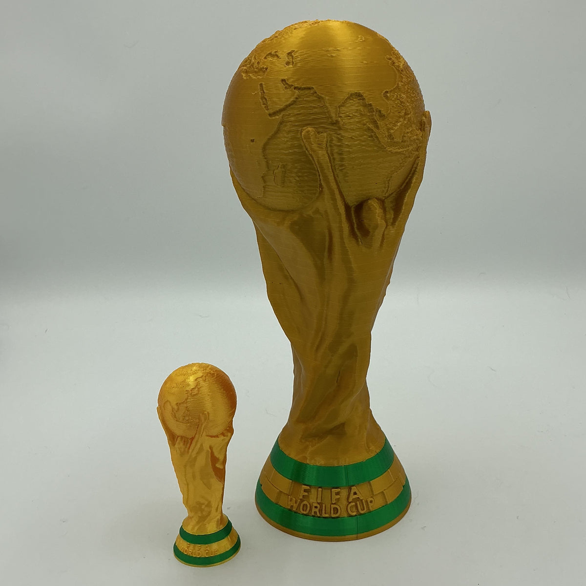 A gold replica of the FIFA World Cup trophy set, featuring a large replica of the official tournament trophy and a smaller miniature version, both with green accents on the base, photographed on a light gray background.