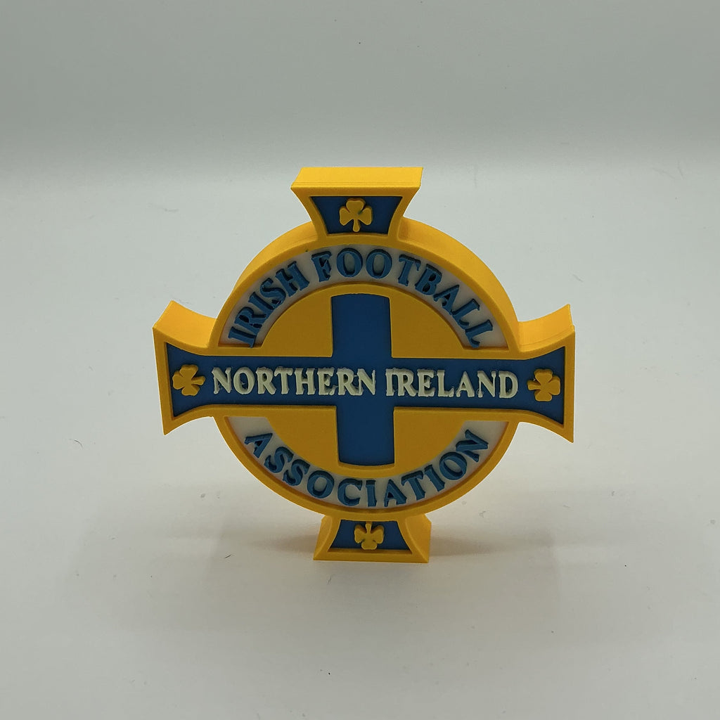 A yellow and blue Northern Ireland Football Association cross-shaped sign or plaque on a light gray studio background.