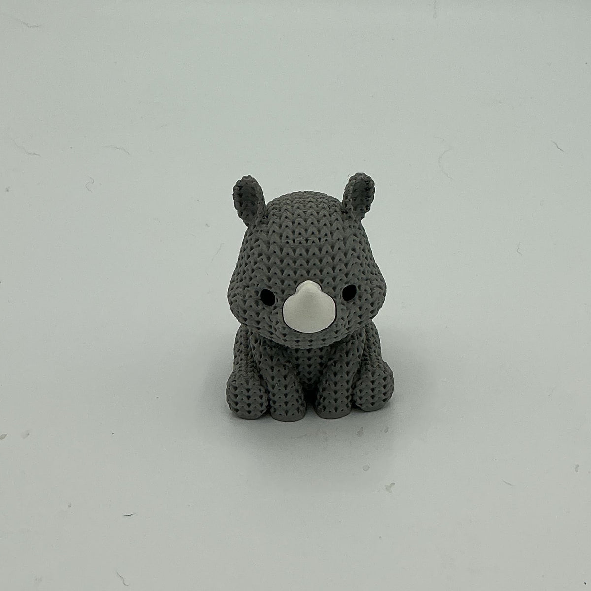 A small gray knitted-style rhino figurine sitting on a plain light gray surface against a matching gray background.