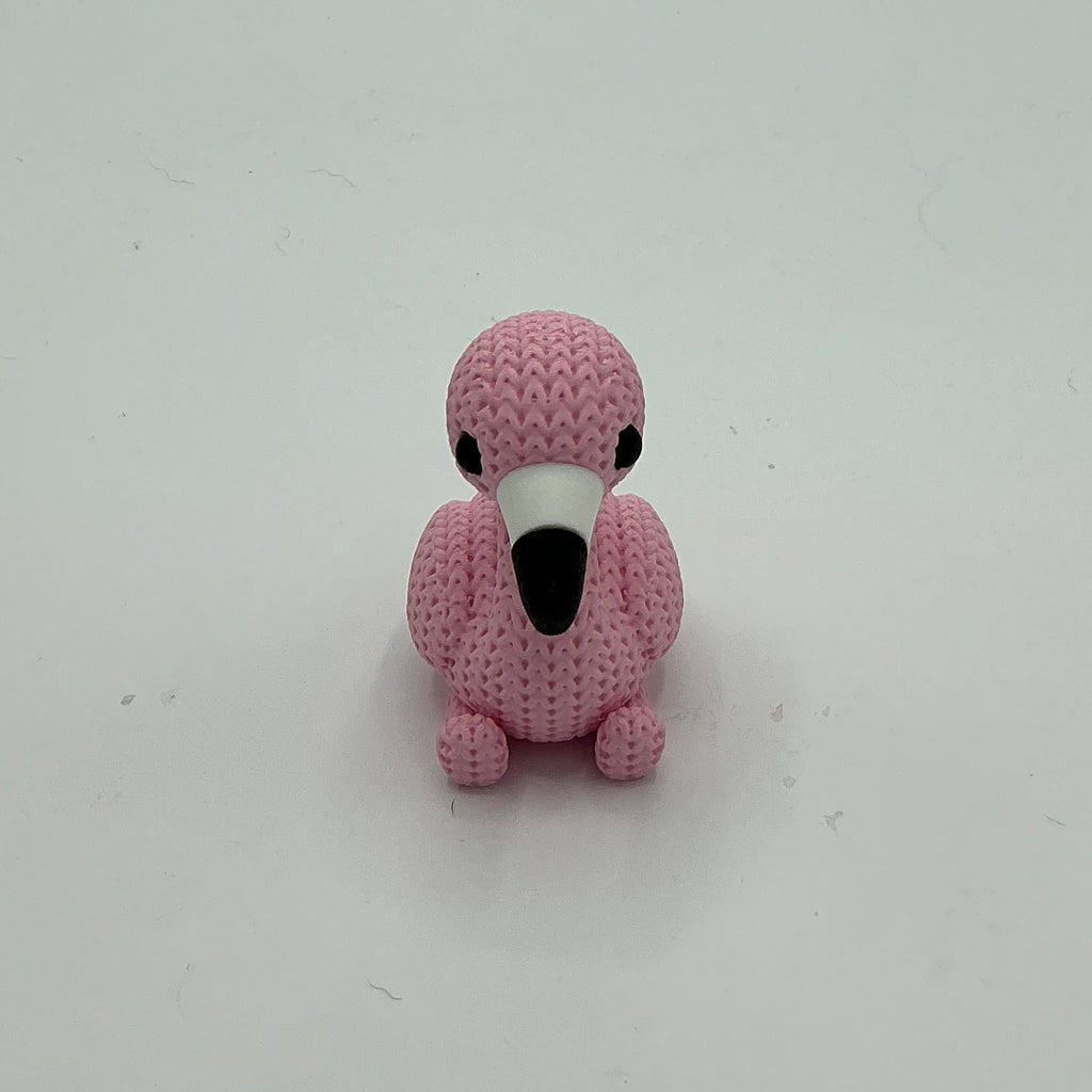 A small pink crocheted-style flamingo figurine with a black beak and eyes, standing on a light gray surface against a matching gray background.