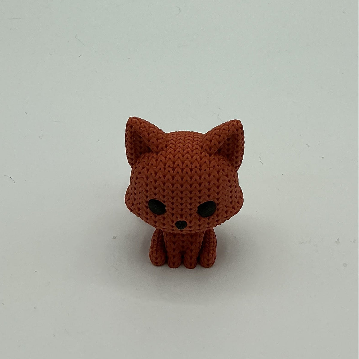 A small red-orange knitted-style fox figurine with black eyes and nose, standing on a light gray background.