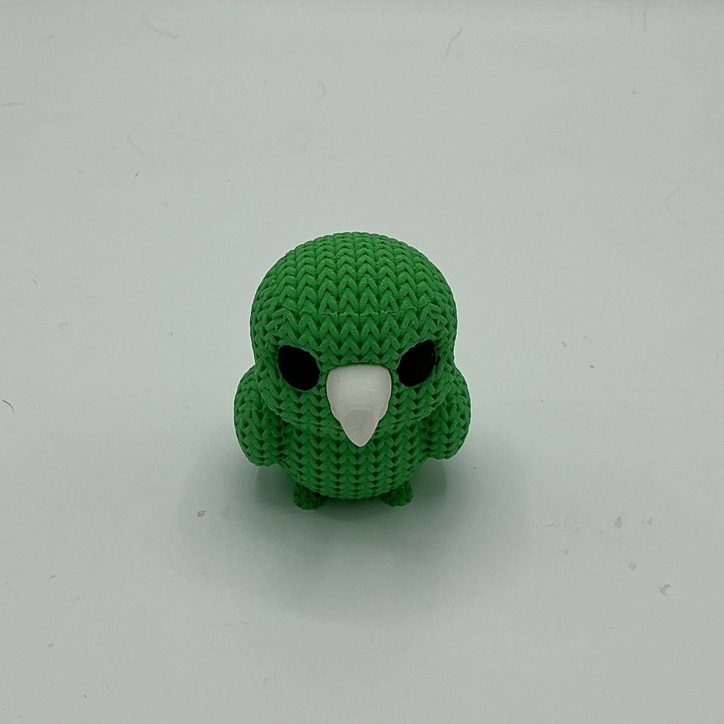 A small green knitted-style bird figurine with a white beak and black eyes, standing upright on a plain light gray background.