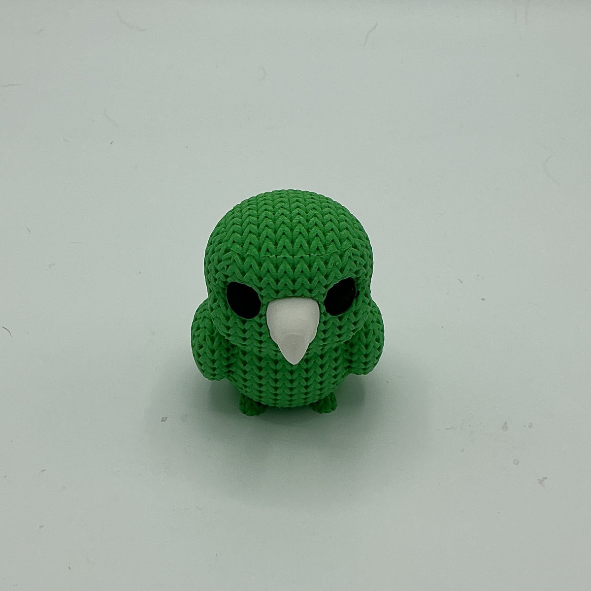 A small green knitted-style bird figurine with a white beak and black eyes, standing upright on a plain light gray background.