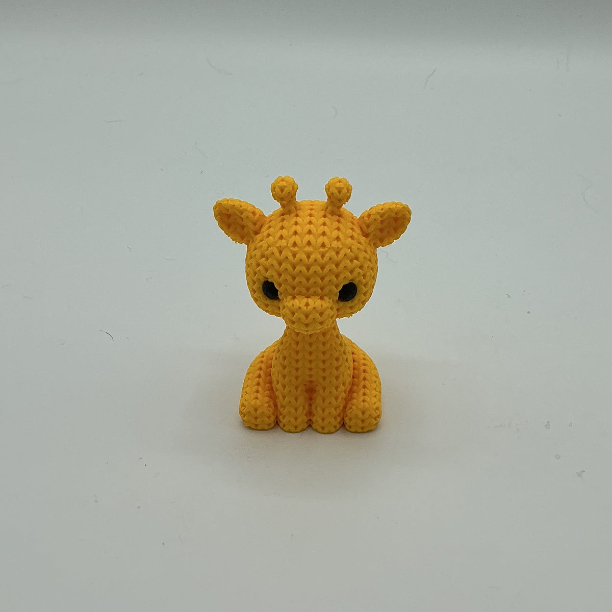 A small crocheted-style giraffe figurine in bright yellow-orange, sitting on a plain light gray studio background.