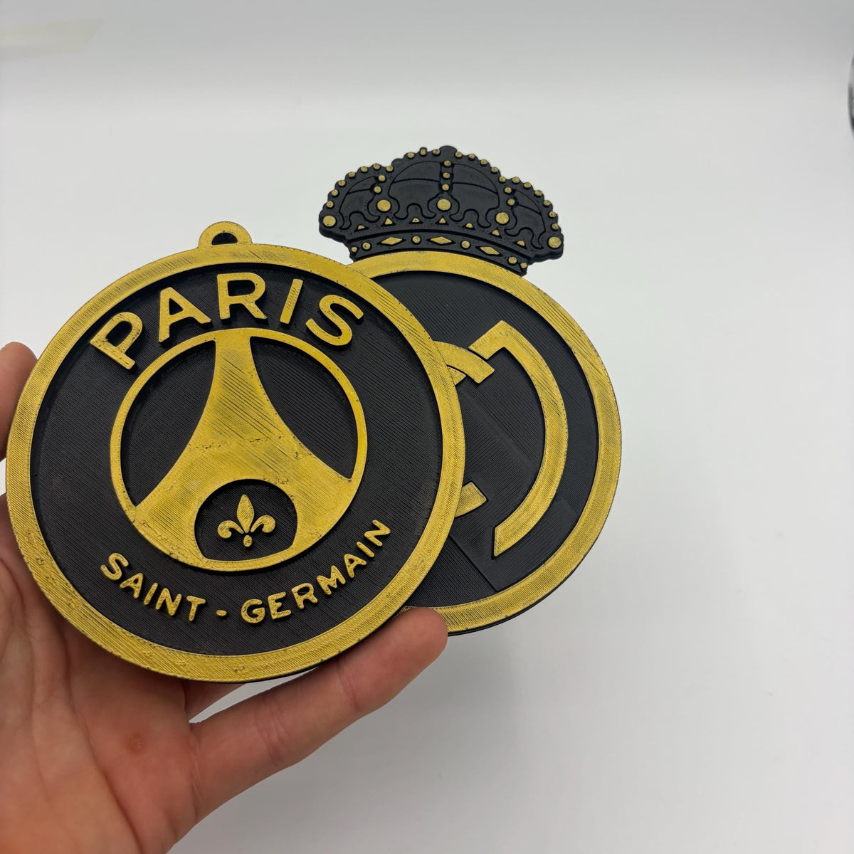A hand holding a Paris Saint-Germain circular wall plaque or ornament, with a second matching plaque partially visible behind it against a plain light background.