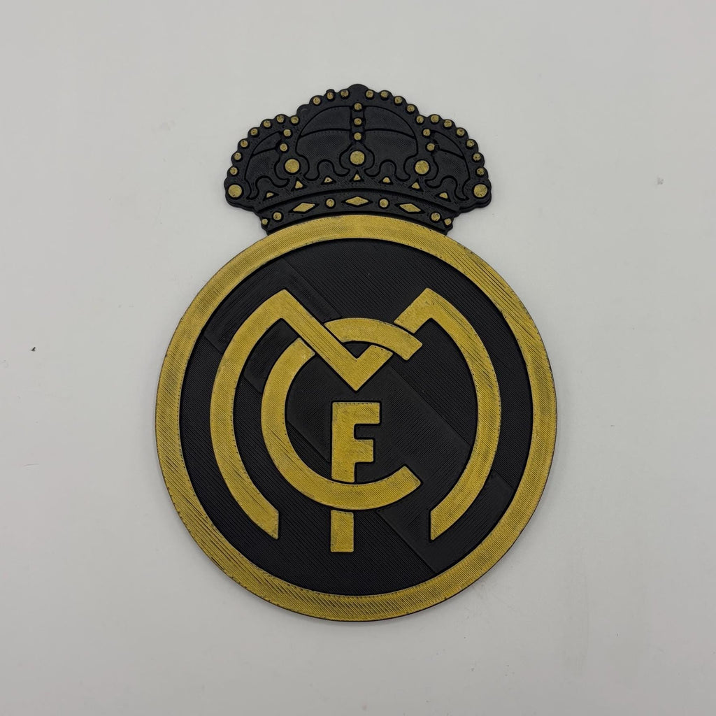 A Real Madrid FC crest emblem with a black and gold color scheme, featuring a crown at the top and the interlocking 'R' and 'F' logo in the center, shown on a plain light background.