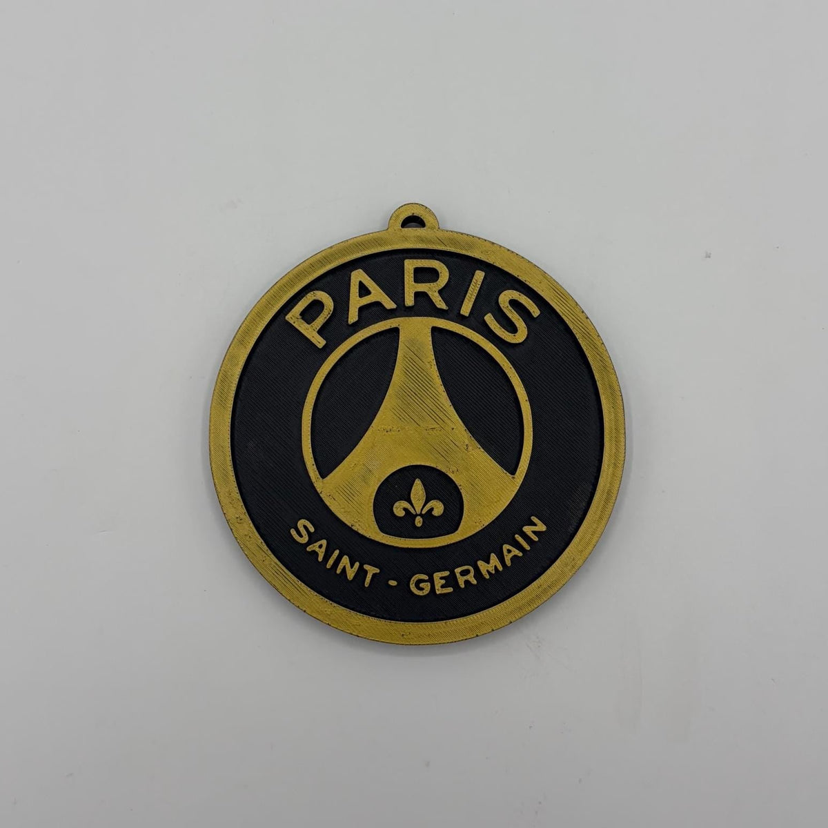 A circular Paris Saint-Germain metal badge or charm with a gold and black design, featuring the club's logo and text, displayed on a plain light gray background.