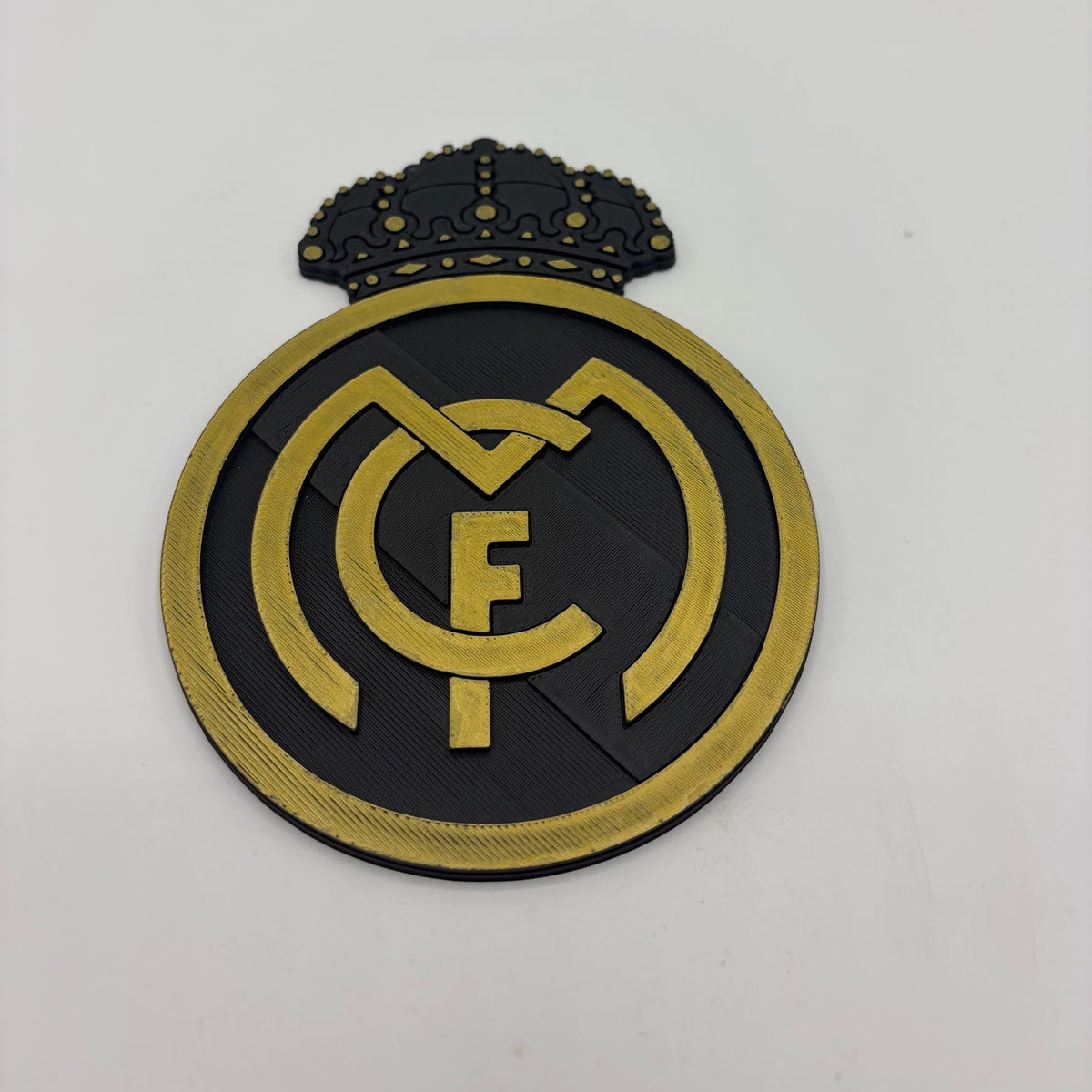 A Real Madrid crest patch or emblem with a black and gold design, featuring the club's initials and a crown on a plain light background.