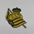 A 3D wooden cutout of the Real Sociedad crest, featuring a crown, soccer ball, and striped flag with a monogram 'S', displayed on a plain light gray background.