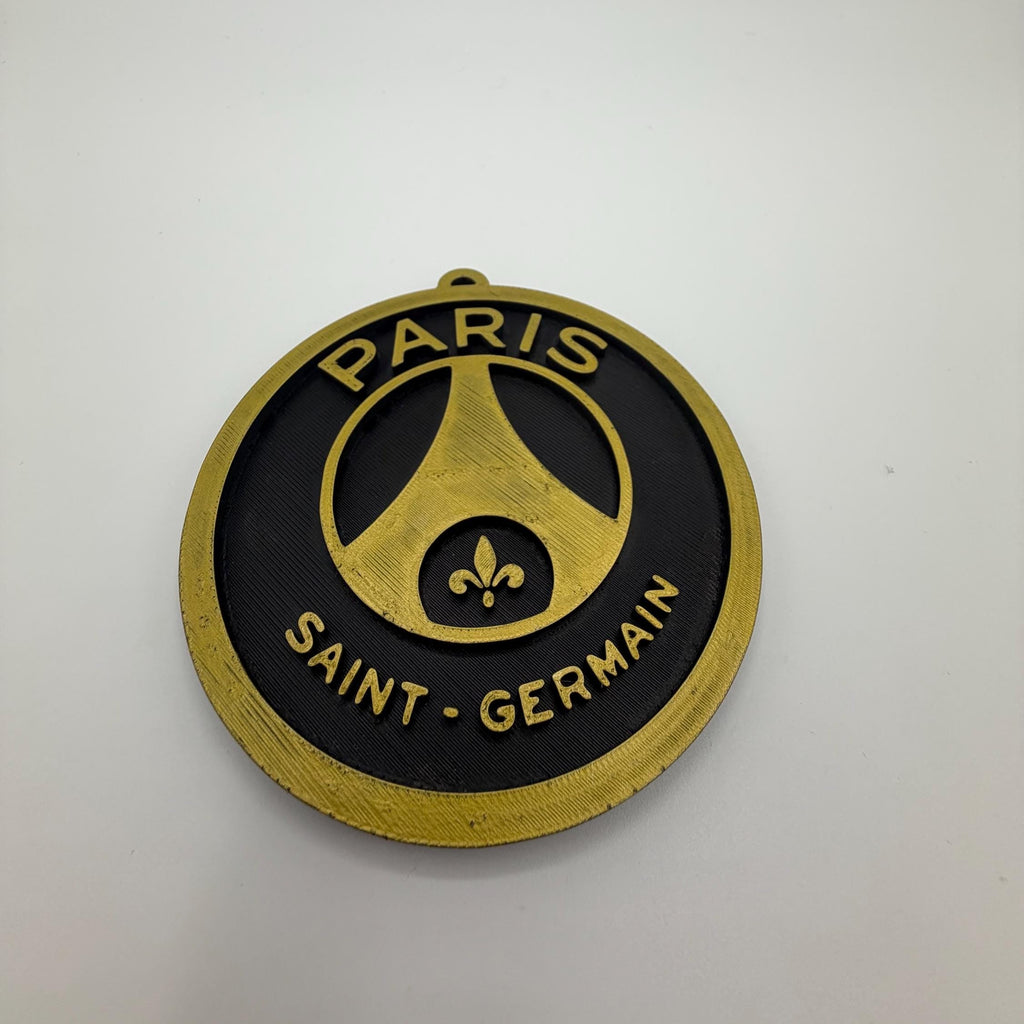 A circular Paris Saint-Germain football club badge or medallion with a black face, gold border, and gold lettering reading 'PARIS' and 'SAINT-GERMAIN', photographed on a plain light background.