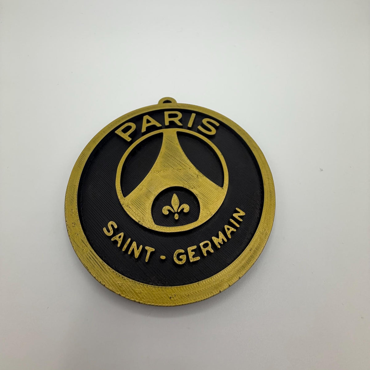 A circular Paris Saint-Germain football club badge or medallion with a black face, gold border, and gold lettering reading 'PARIS' and 'SAINT-GERMAIN', photographed on a plain light background.