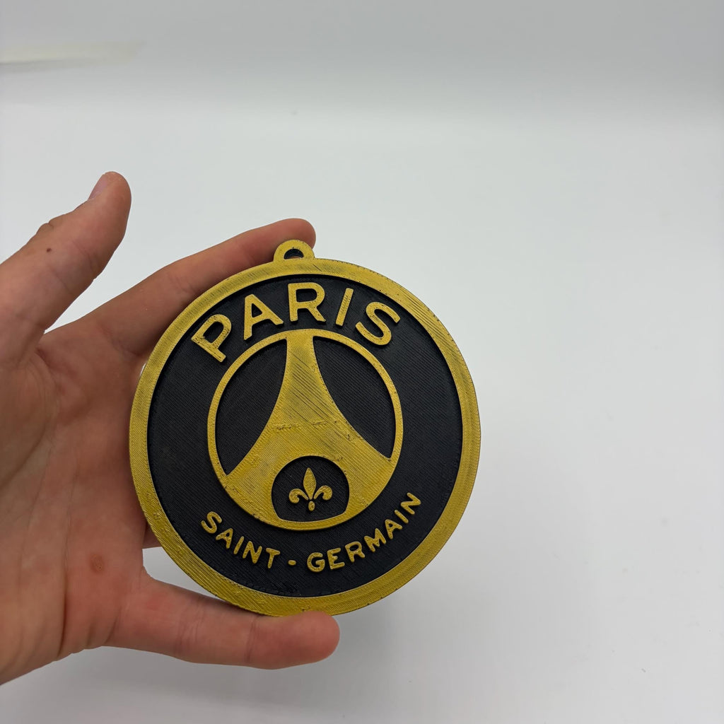 A hand holding a round Paris Saint-Germain FC wooden plaque or ornament against a plain white background.