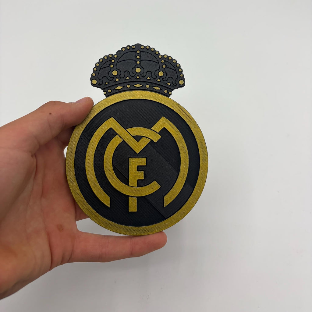 A hand holding a circular black and gold Real Madrid crest plaque with a crown on top, set against a plain white background.