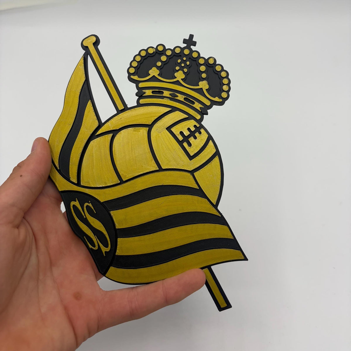 A hand holding a yellow and black cutout of the Real Sociedad soccer club crest, featuring a crown, soccer ball, and striped flag, against a plain white background.