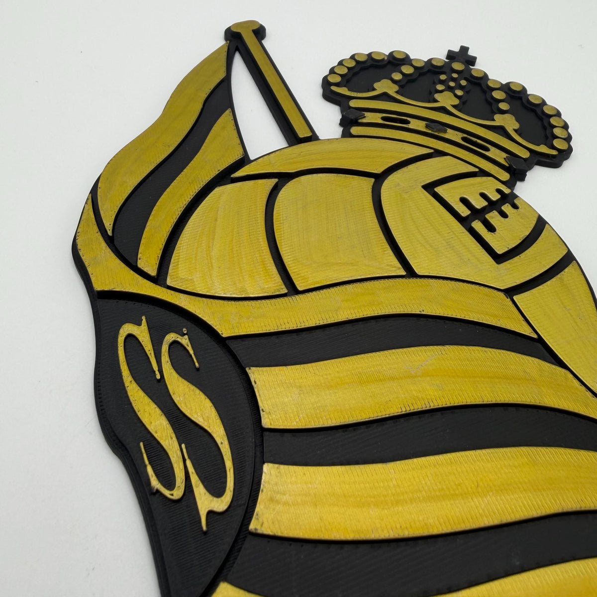 A close-up of a yellow and black striped soccer ball crest with a crown and stylized 'SS' emblem, photographed against a plain light background.
