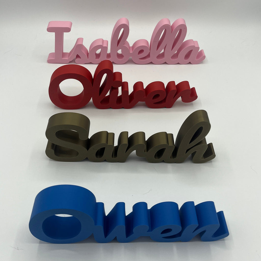 Four personalized name signs spelling Isabella, Oliver, Sarah, and Owen in pink, red, bronze, and blue, arranged vertically on a white surface.