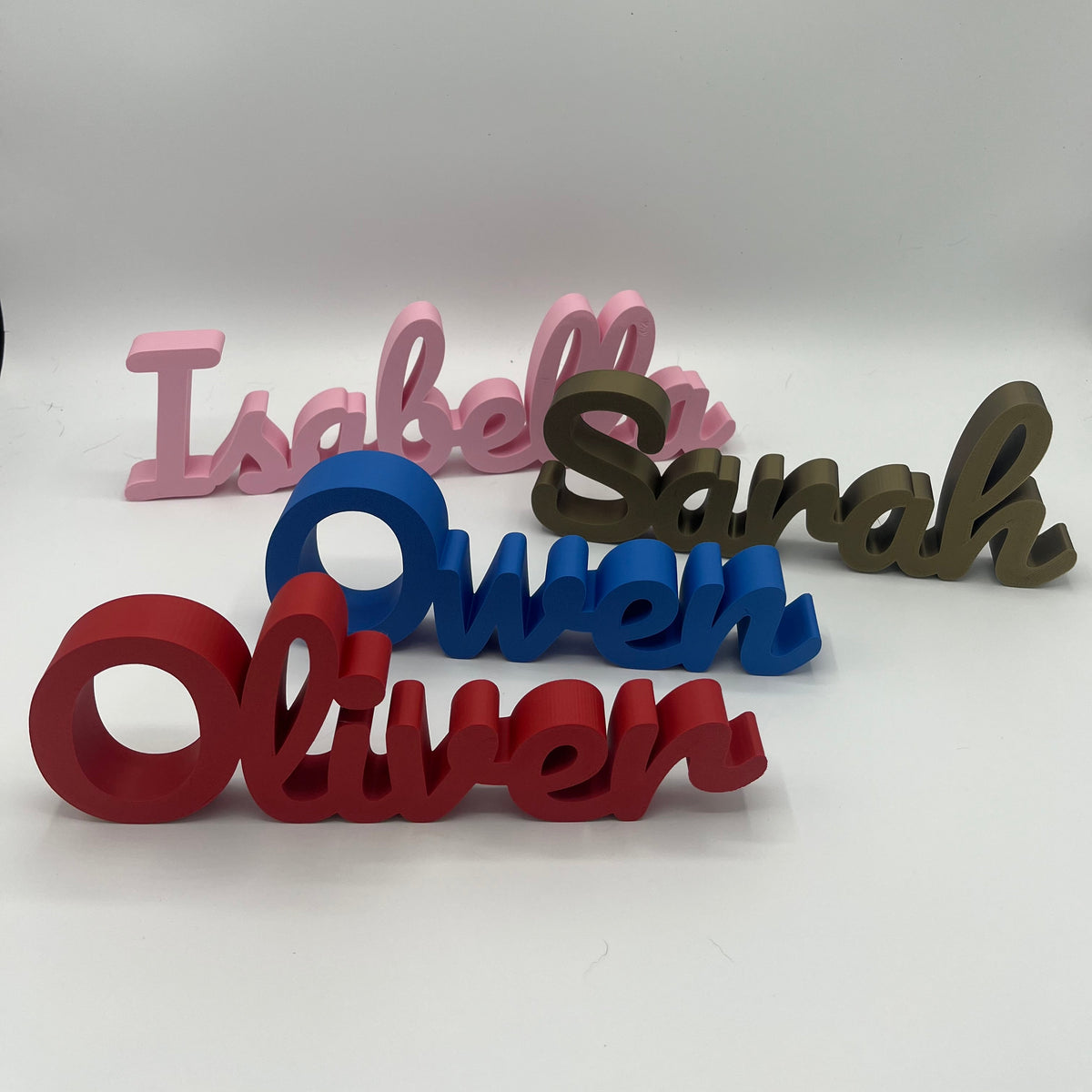 Five 3D custom name signs in pink, blue, red, and bronze, spelling out the names Isabella, Sarah, Owen, and Oliver, arranged on a light grey background.
