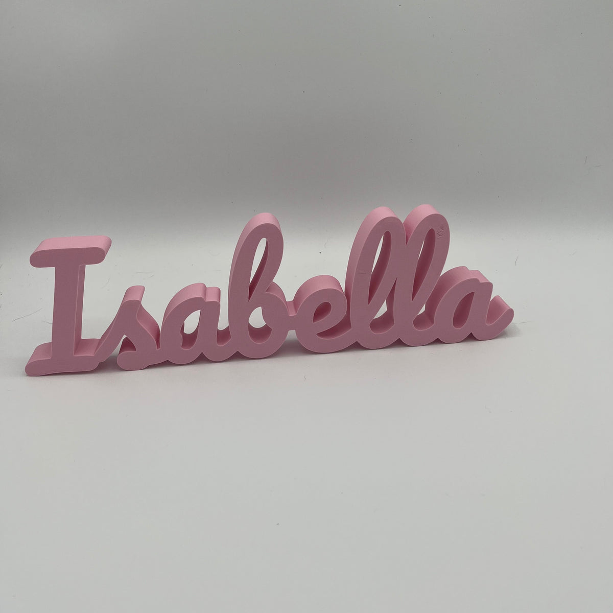 A pink 3D printed name sign spelling 'Isabella' in a cursive font, shown on a neutral gray background.