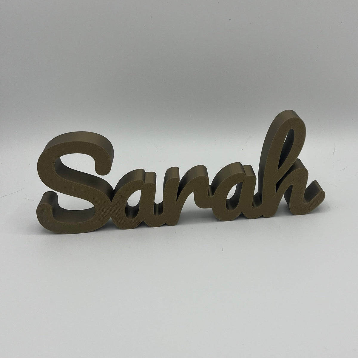 A 3D printed name sign spelling 'Sarah' in a cursive font, finished in a bronze tone, set against a plain light grey background.