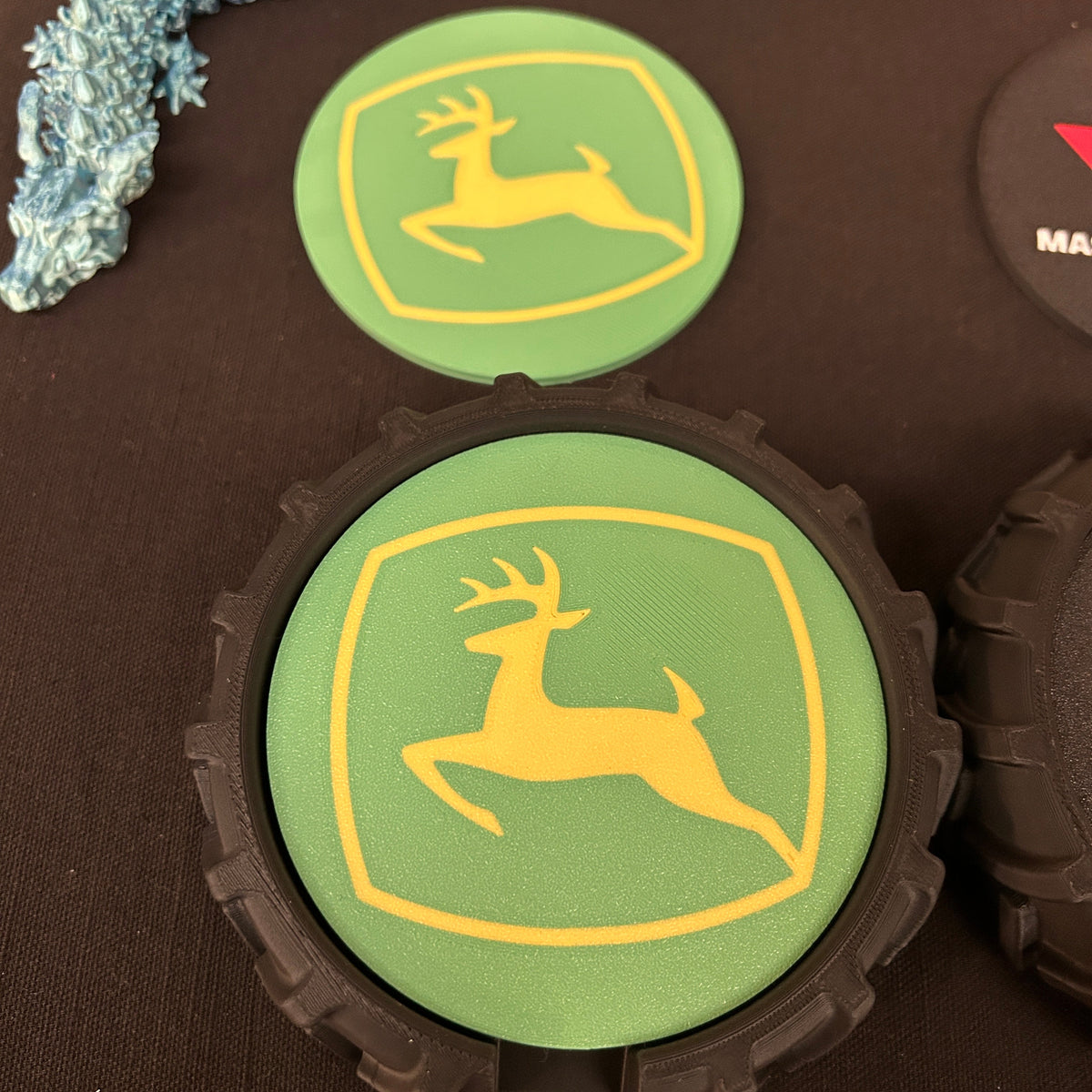 A close-up of a John Deere-themed coaster or decal featuring the iconic green and yellow deer logo, shown on a dark textured surface with other similar items partially visible.