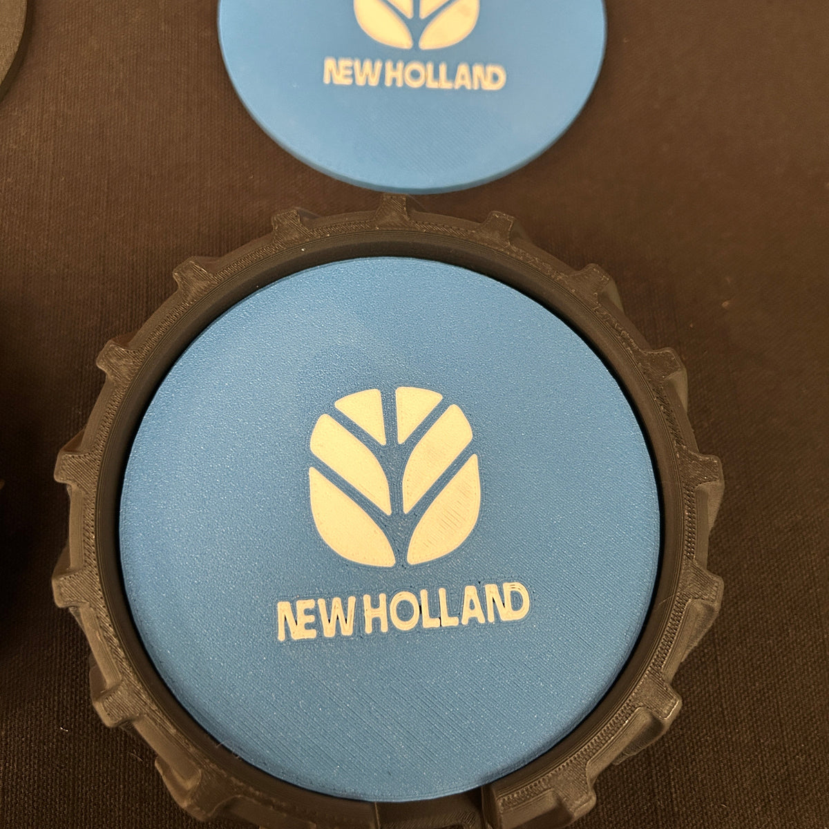 A close-up of a circular blue New Holland branded item with a black rugged outer rim, placed on a dark brown textured surface. A second matching blue circular piece is visible above it.