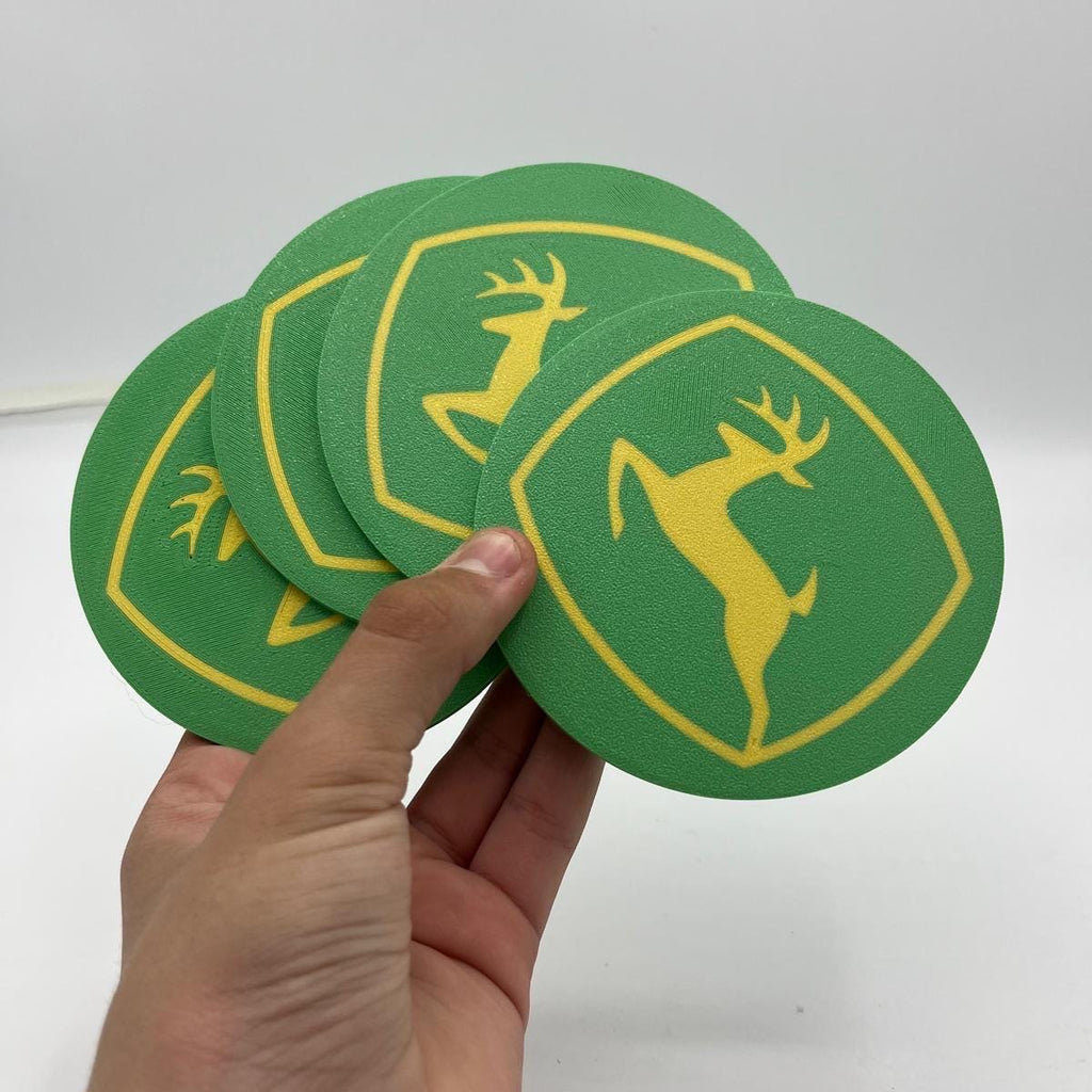 A hand holding a stack of green circular coasters with a yellow outline and a yellow deer logo, shown against a plain white background.