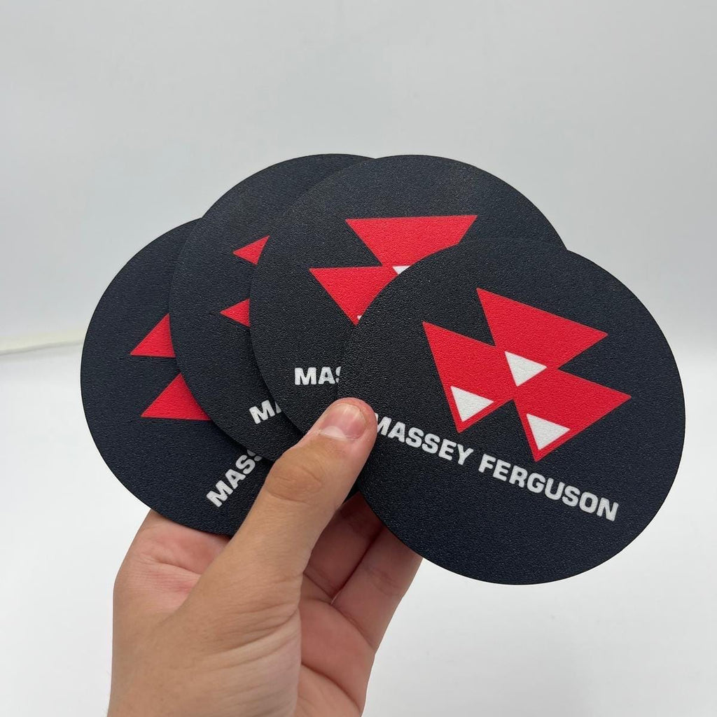 A hand holds four round black coasters featuring a red geometric logo and the name MASSEY FERGUSON in white text, against a plain light background.