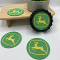 Three green John Deere coasters with yellow logos, including one round coaster and two oval coasters, displayed on a white surface with a small wooden stand and a potted plant in the background.