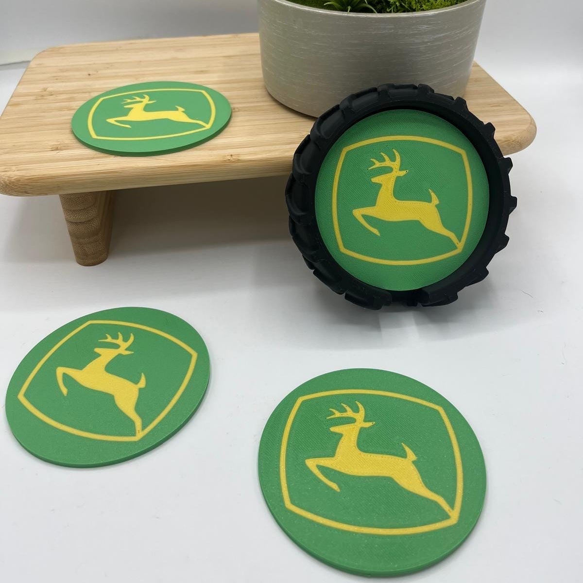 Three green John Deere coasters with yellow logos, including one round coaster and two oval coasters, displayed on a white surface with a small wooden stand and a potted plant in the background.