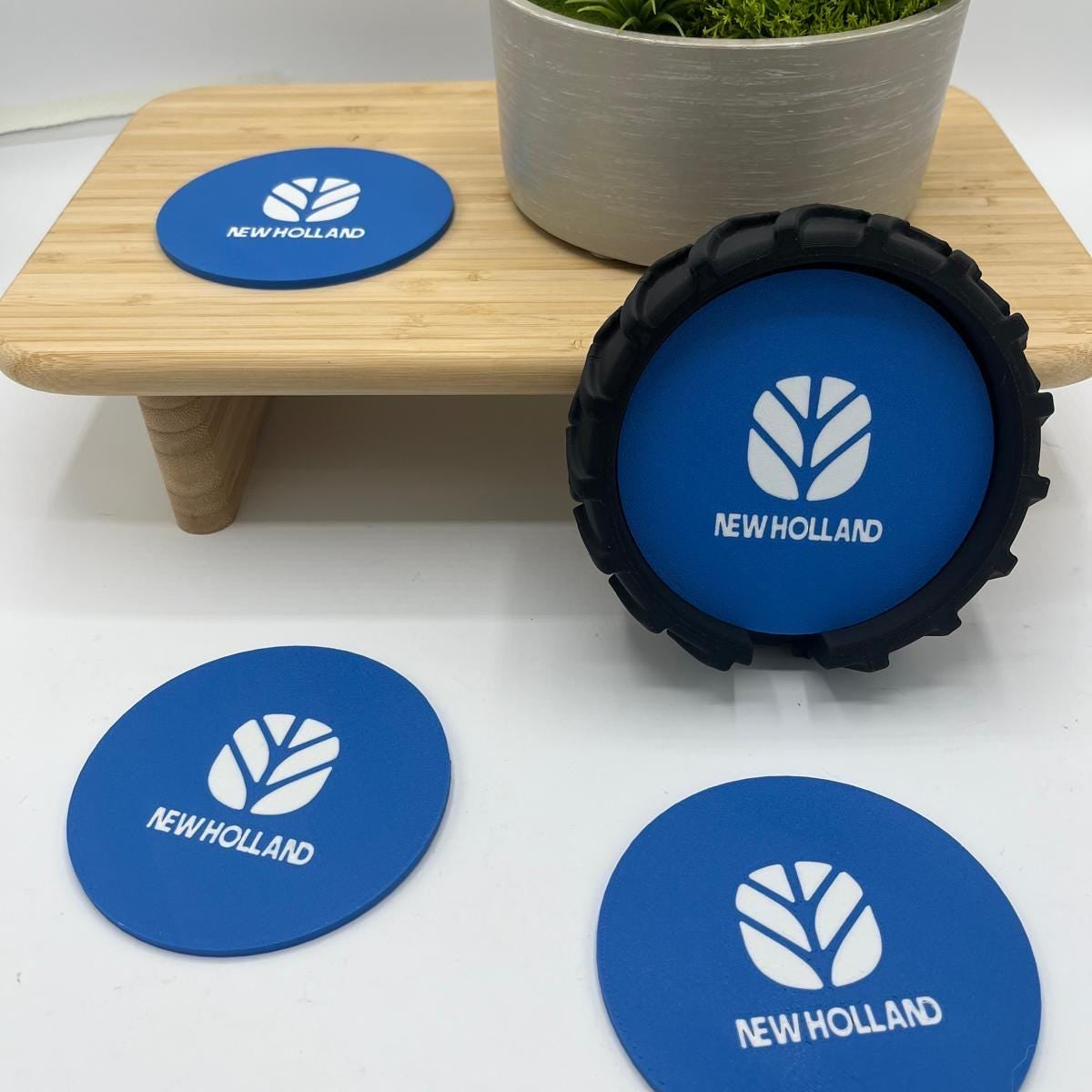 Three blue circular New Holland branded coasters are arranged on a white surface, with one standing upright in a black textured holder and two lying flat. A small wooden riser and a potted green plant sit behind them.