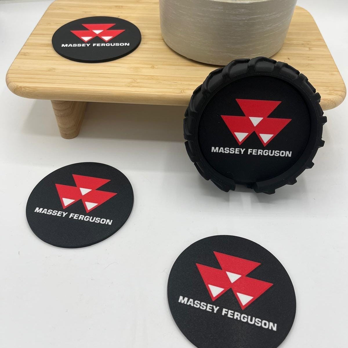 Three black circular coasters with a red and white geometric logo and the text 'MASSEY FERGUSON' are displayed on a white surface, with one coaster standing upright on a small wooden riser next to a potted green plant.