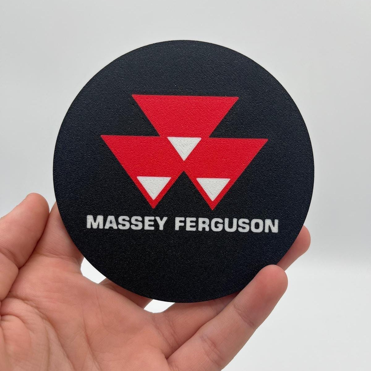 A hand holding a round black coaster featuring a red geometric logo and the text 'MASSEY FERGUSON' in white, against a plain light gray background.