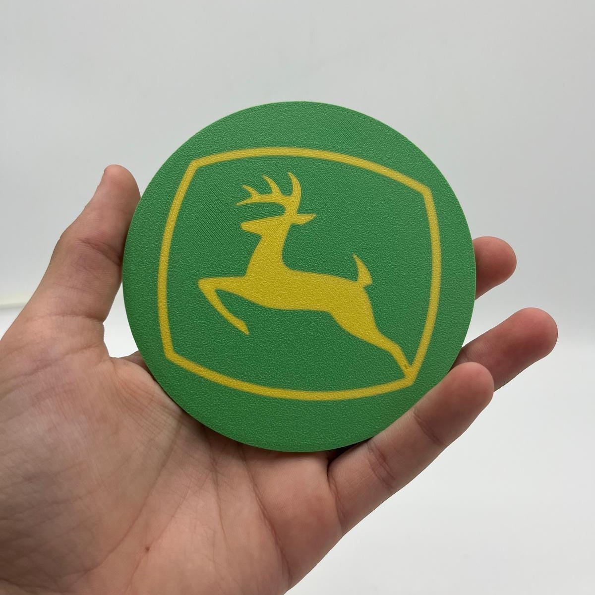 A hand holding a round green and yellow John Deere coaster against a plain light gray background.