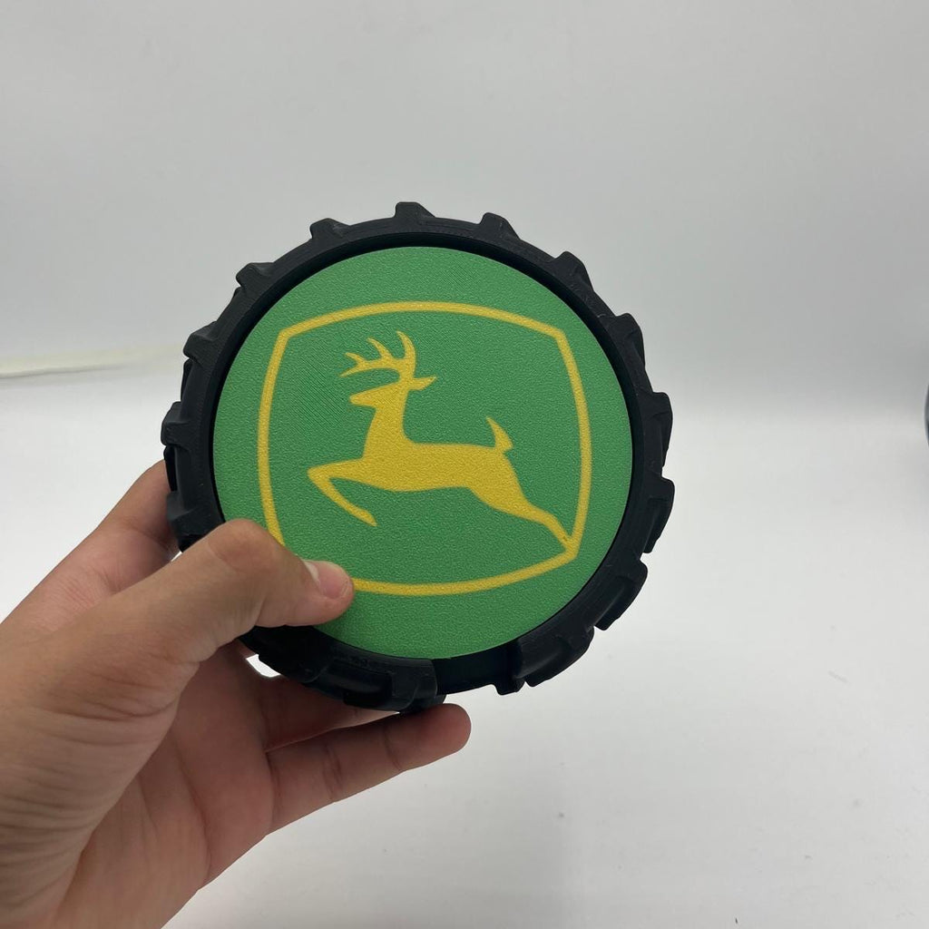 A hand holding a circular black tyre featuring the green and yellow John Deere logo coasters on a white background.