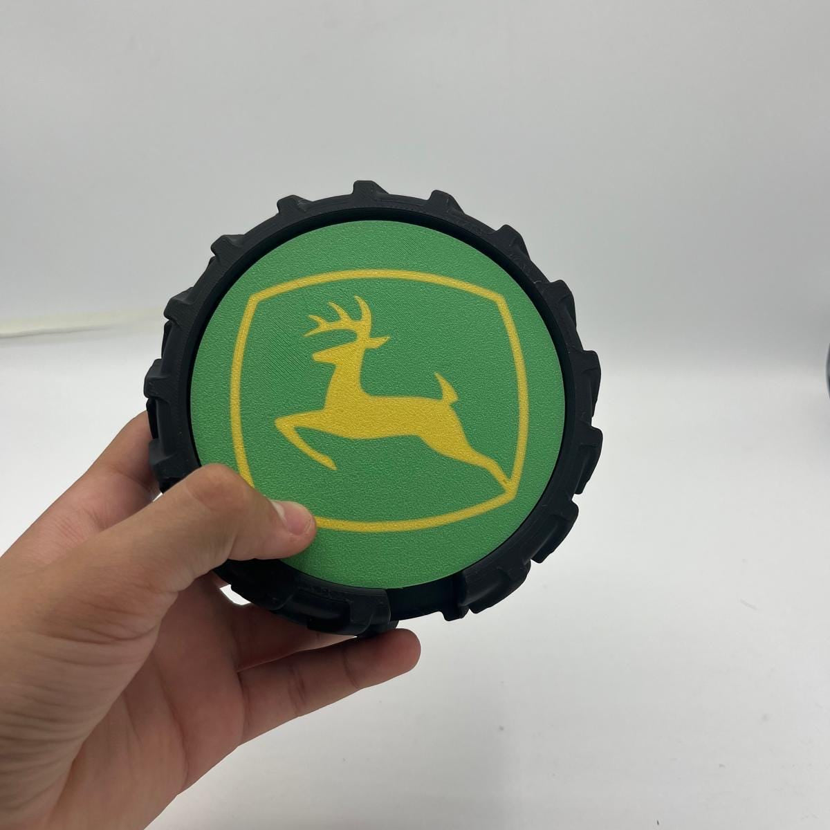 A hand holding a circular black tyre featuring the green and yellow John Deere logo coasters on a white background.