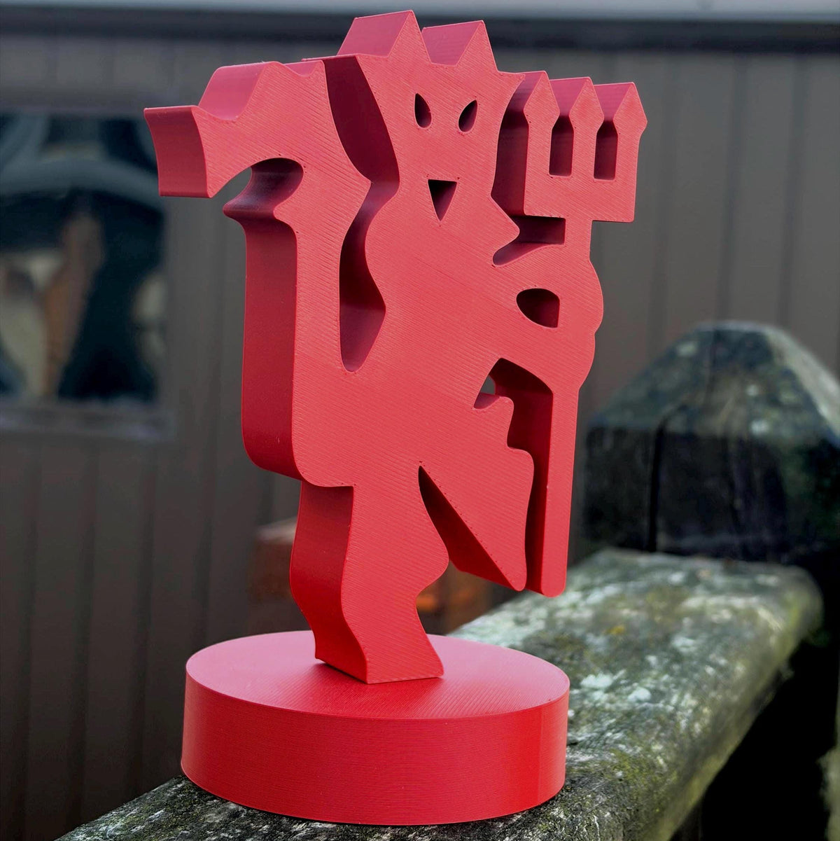 A red 3D-printed decorative sculpture of a stylised devil rampant holding a trident, mounted on a matching round base, photographed outdoors on a weathered wooden railing.